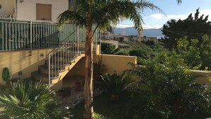Edificio residencial con una escalera que conduce a un balcón, rodeado de abundante vegetación y vistas a un paisaje costero.