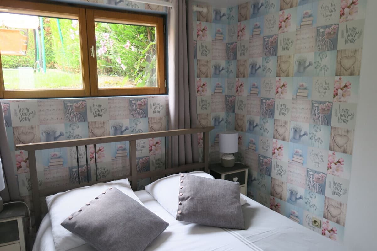 Un dormitorio con cama, dos almohadas, una mesita de noche con lámpara y una ventana con vista a follaje.