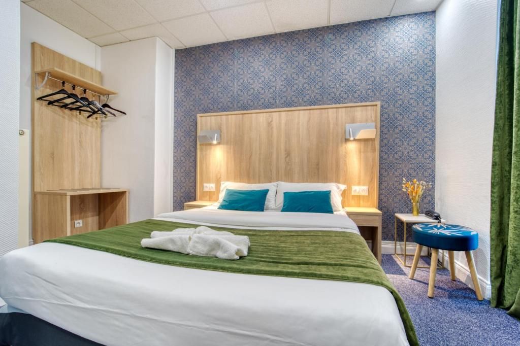 A hotel room with a bed, a wooden headboard, a nightstand, a small table with a vase, and a green curtain.