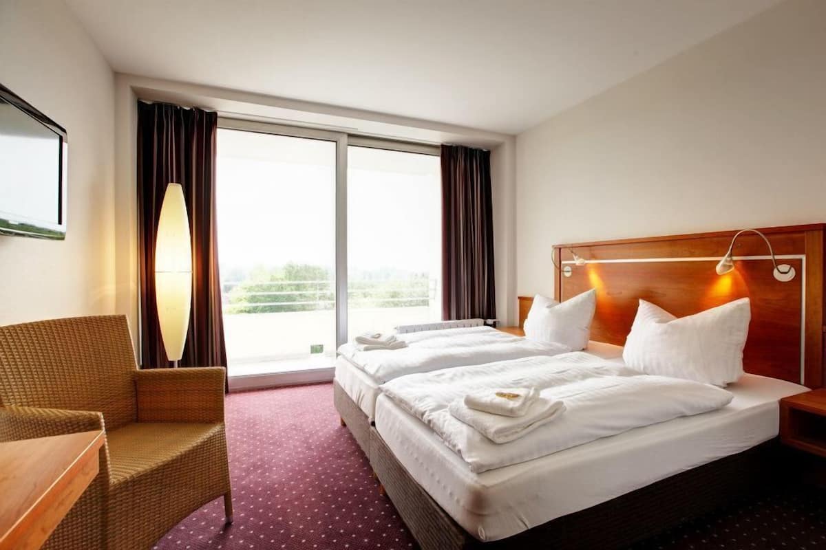 Habitación de hotel con dos camas, un ventanal grande con cortinas, una silla de mimbre y un cabecero de madera con luces.
