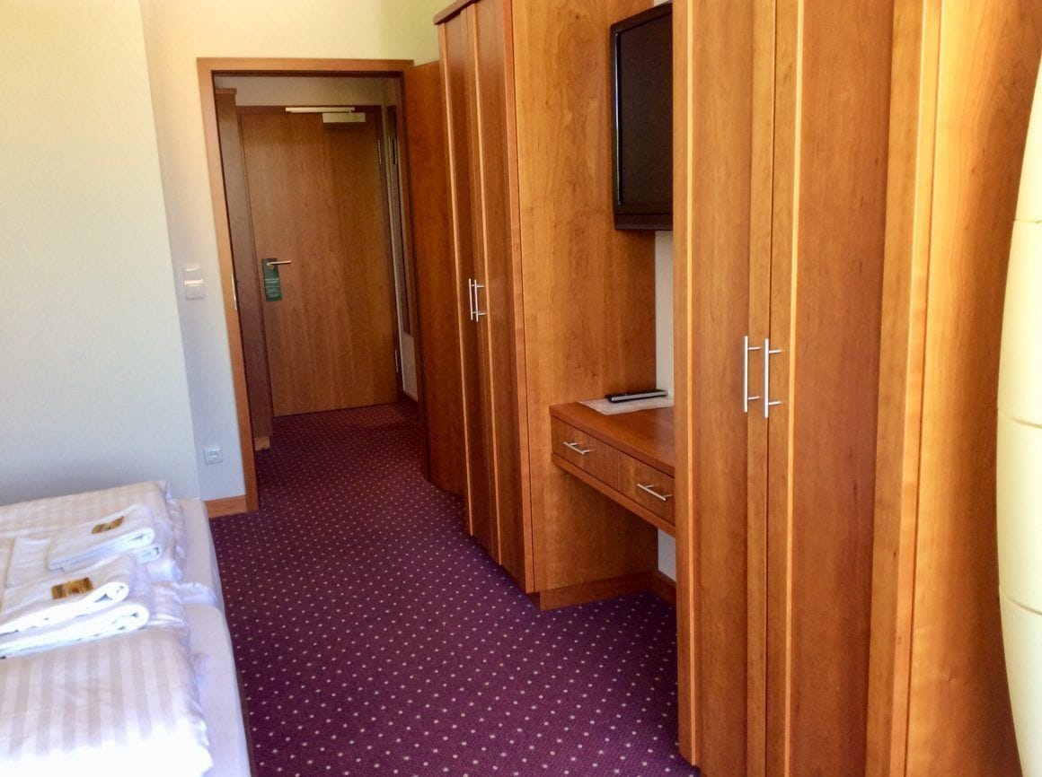 A hotel room with a bed, a wooden wardrobe, a desk with a chair, and a television on the wardrobe.
