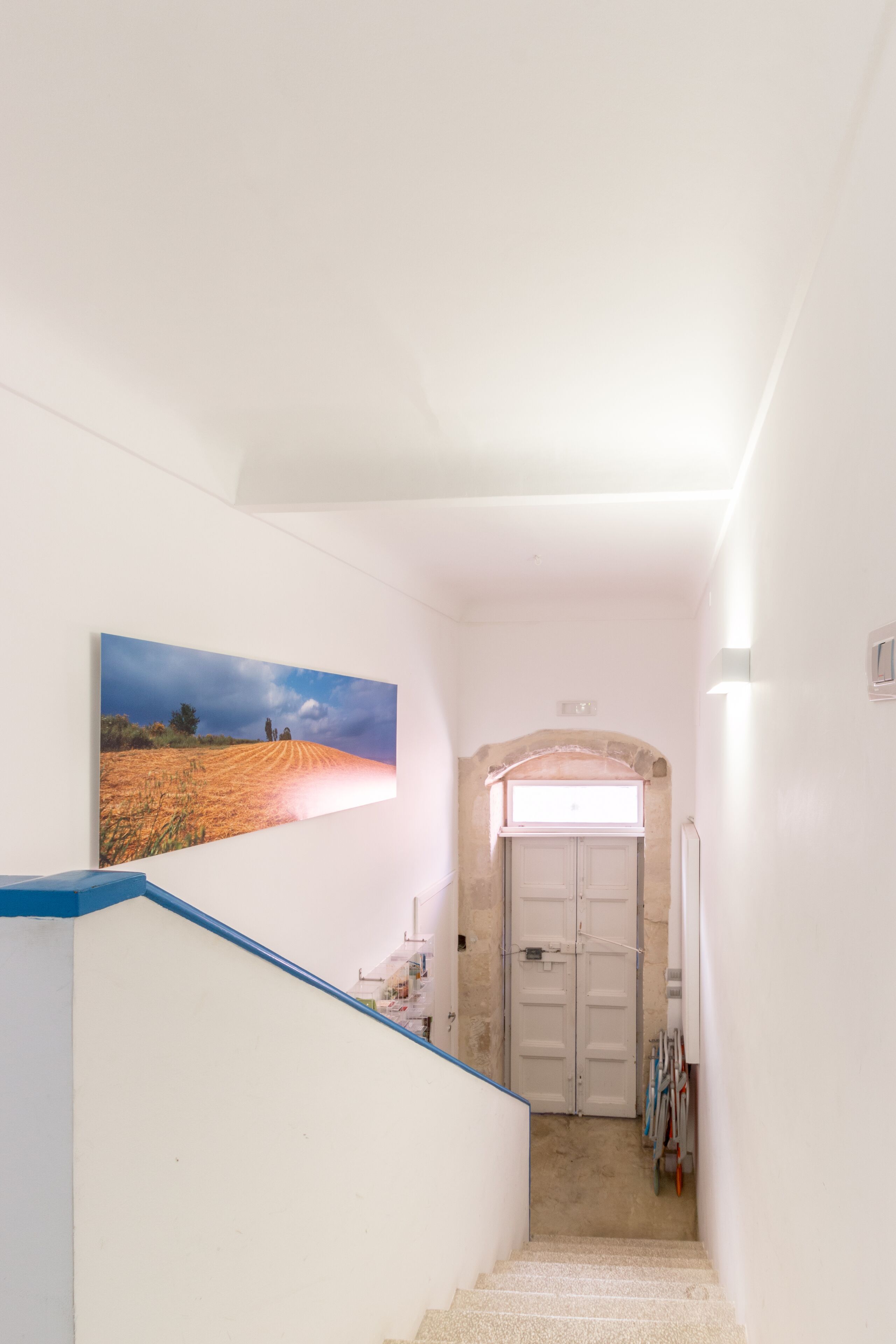 En smal hall med en trappa som leder till en dörr, en inramad landskapsmålning på väggen och ett litet fönster ovanför dörren.