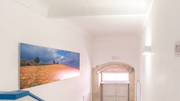 A narrow hallway with a staircase leading to a door, a framed landscape painting on the wall, and a small window above the door.