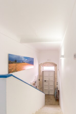 Um corredor estreito com uma escada que leva a uma porta, um quadro de paisagem emoldurado na parede e uma pequena janela acima da porta.