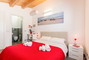 Quarto de hotel com uma cama grande, um quadro com barcos numa praia, uma mesa de cabeceira com abajur vermelho e um guarda-roupa branco.