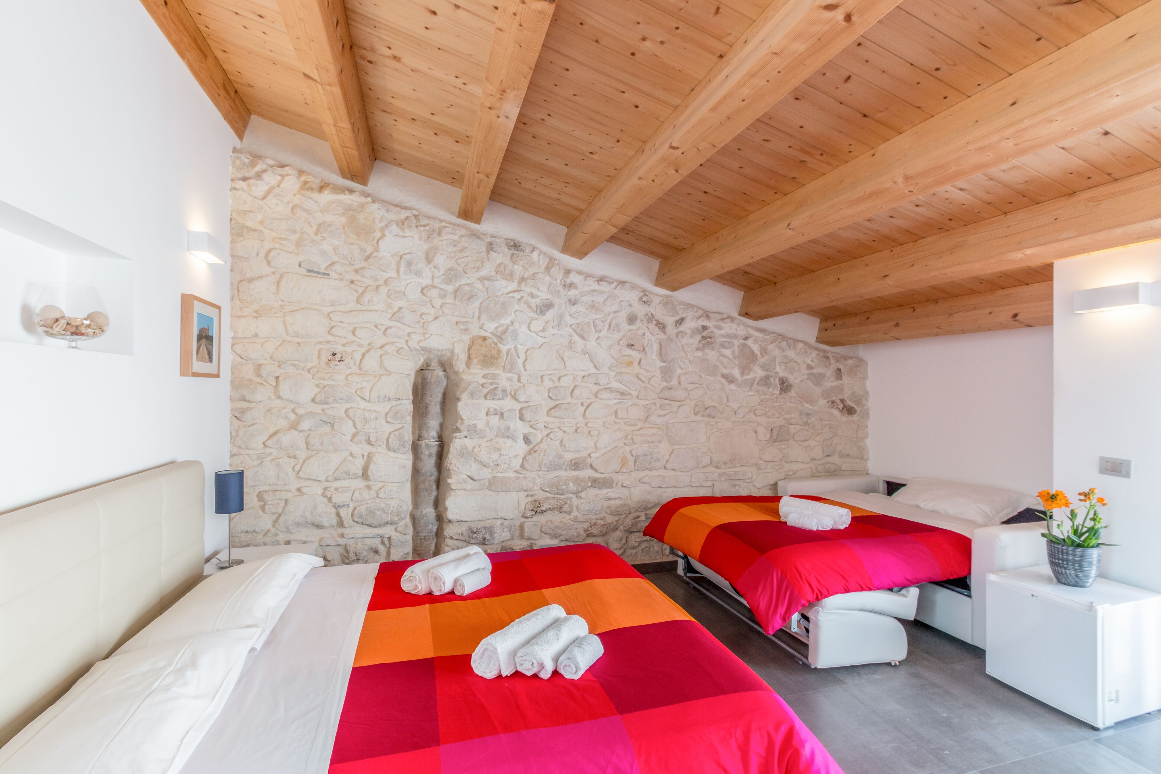 A bedroom with two beds, stone walls, wooden beams, and a small white cabinet with a plant.