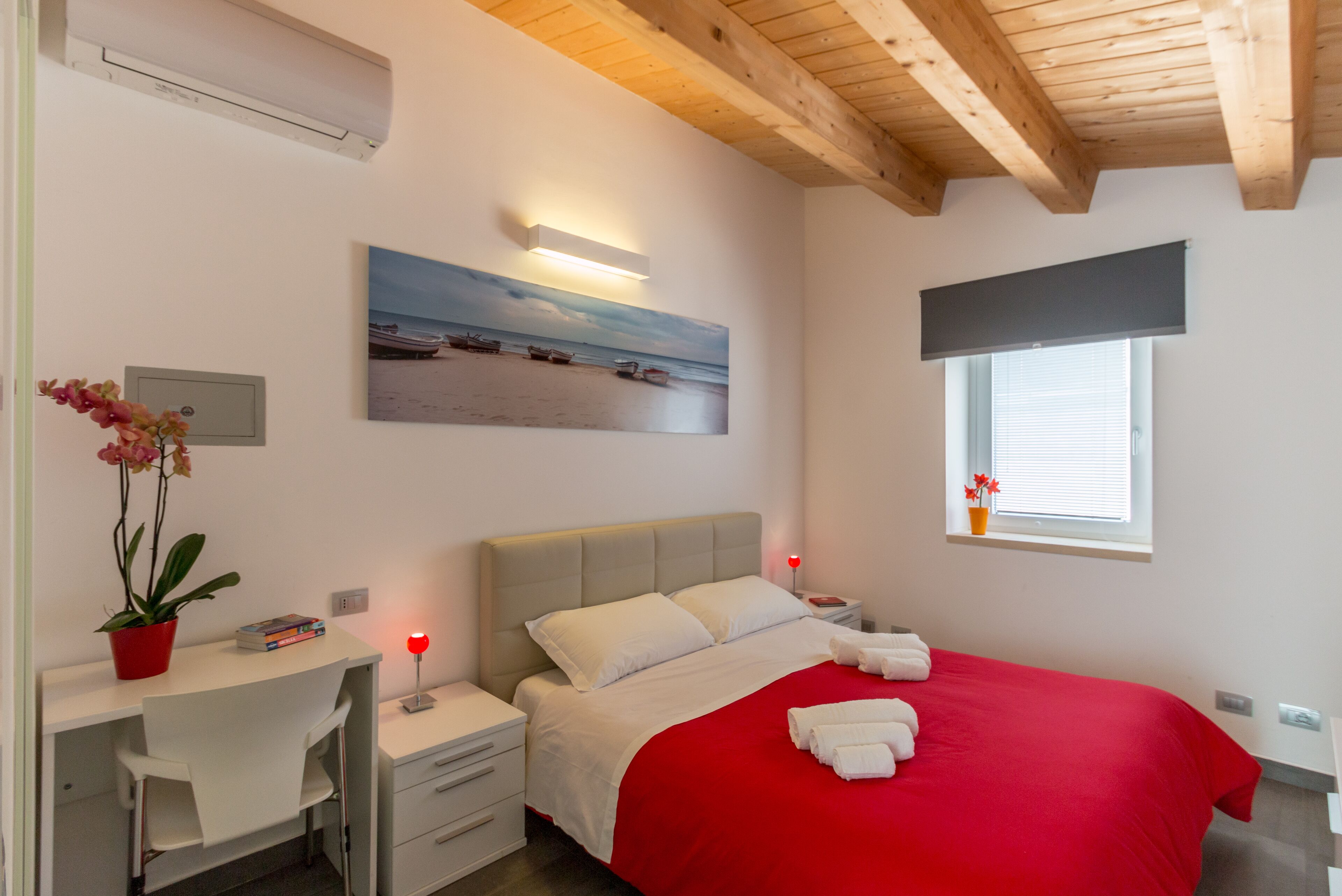 Un dormitorio con cama, escritorio, silla, mesita de noche, ventana y un cuadro en la pared.