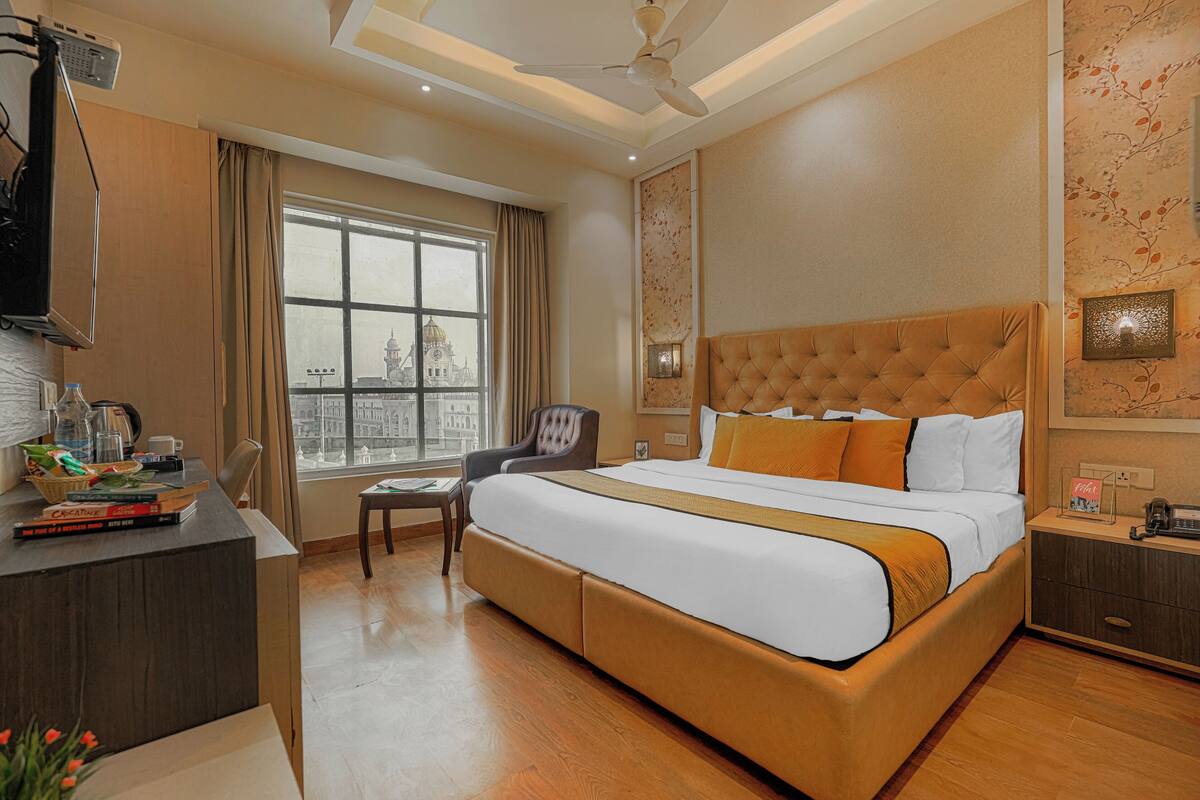 A hotel room with a large bed, a desk with a computer, a chair, a window with a city view, and a ceiling fan.