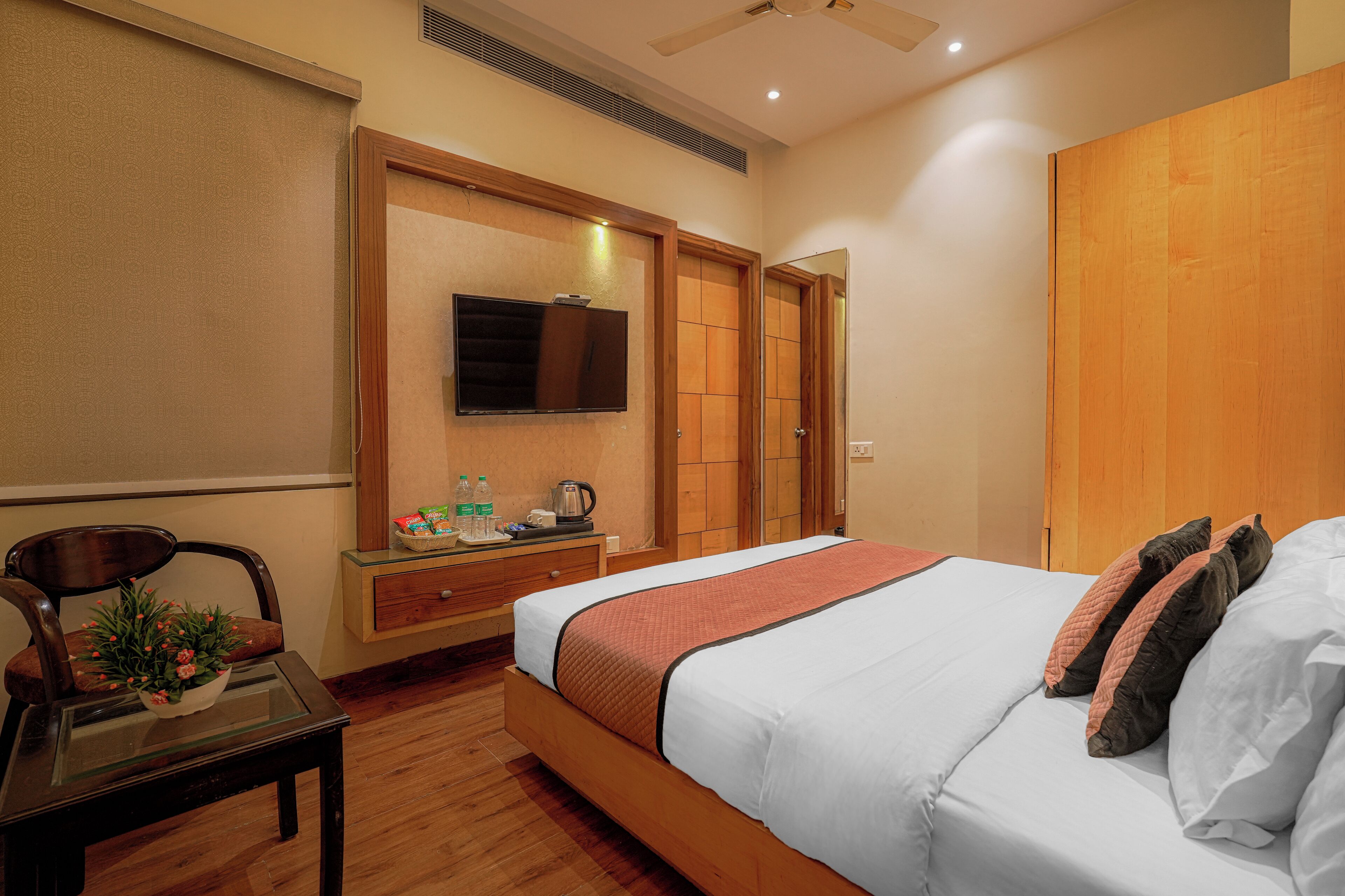 Habitación de hotel con cama, televisor montado en la pared, armario de madera, una pequeña mesa con una planta y un escritorio con silla.