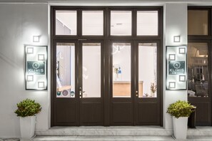 Entrada de hotel con una puerta de madera oscura, dos plantas en macetas y letreros iluminados a ambos lados.