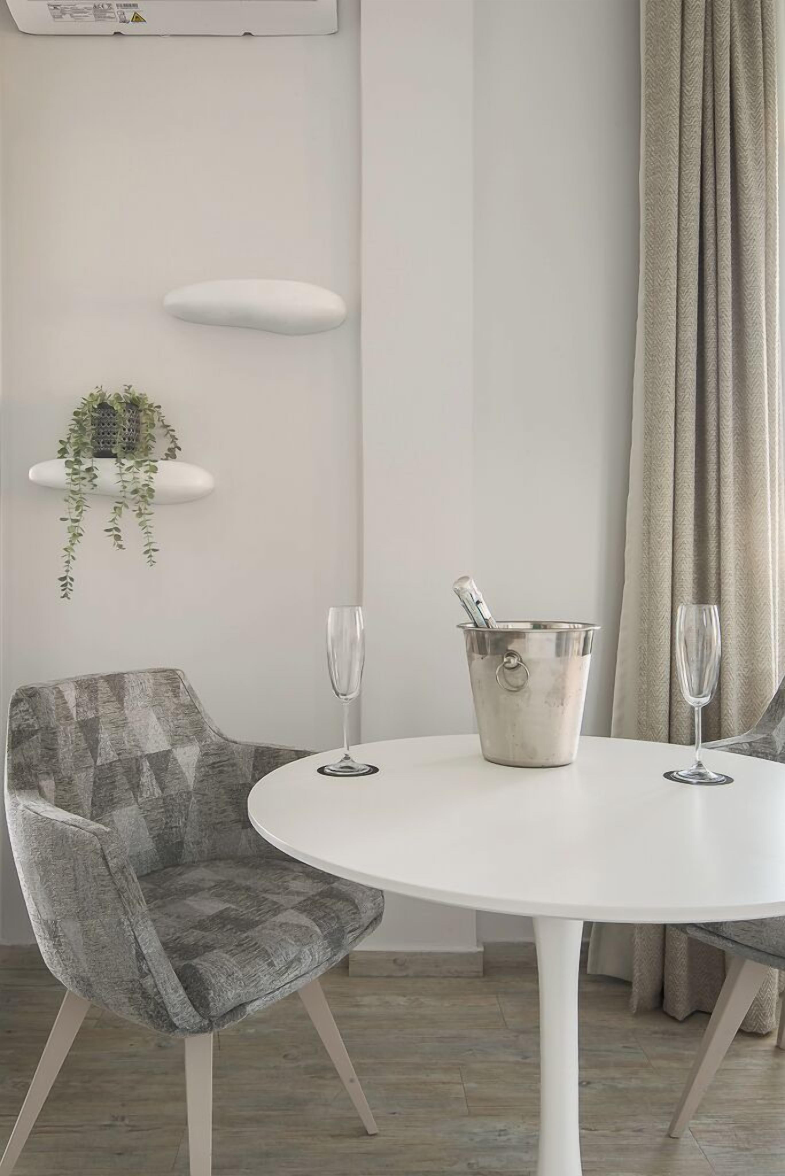 A modern dining area with a white round table, two grey upholstered chairs, a metal bucket with a bottle, and two empty wine glasses.