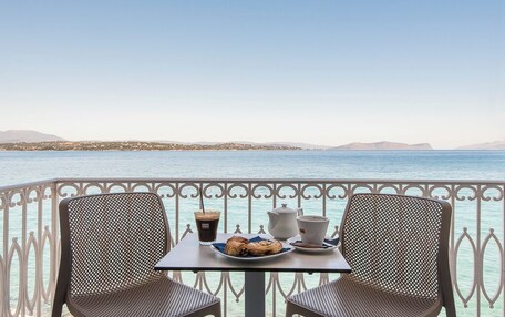 A balcony with two chairs, a table with a plate of pastries, a cup of coffee, and a teapot, overlooking a calm sea and distant mountains.
