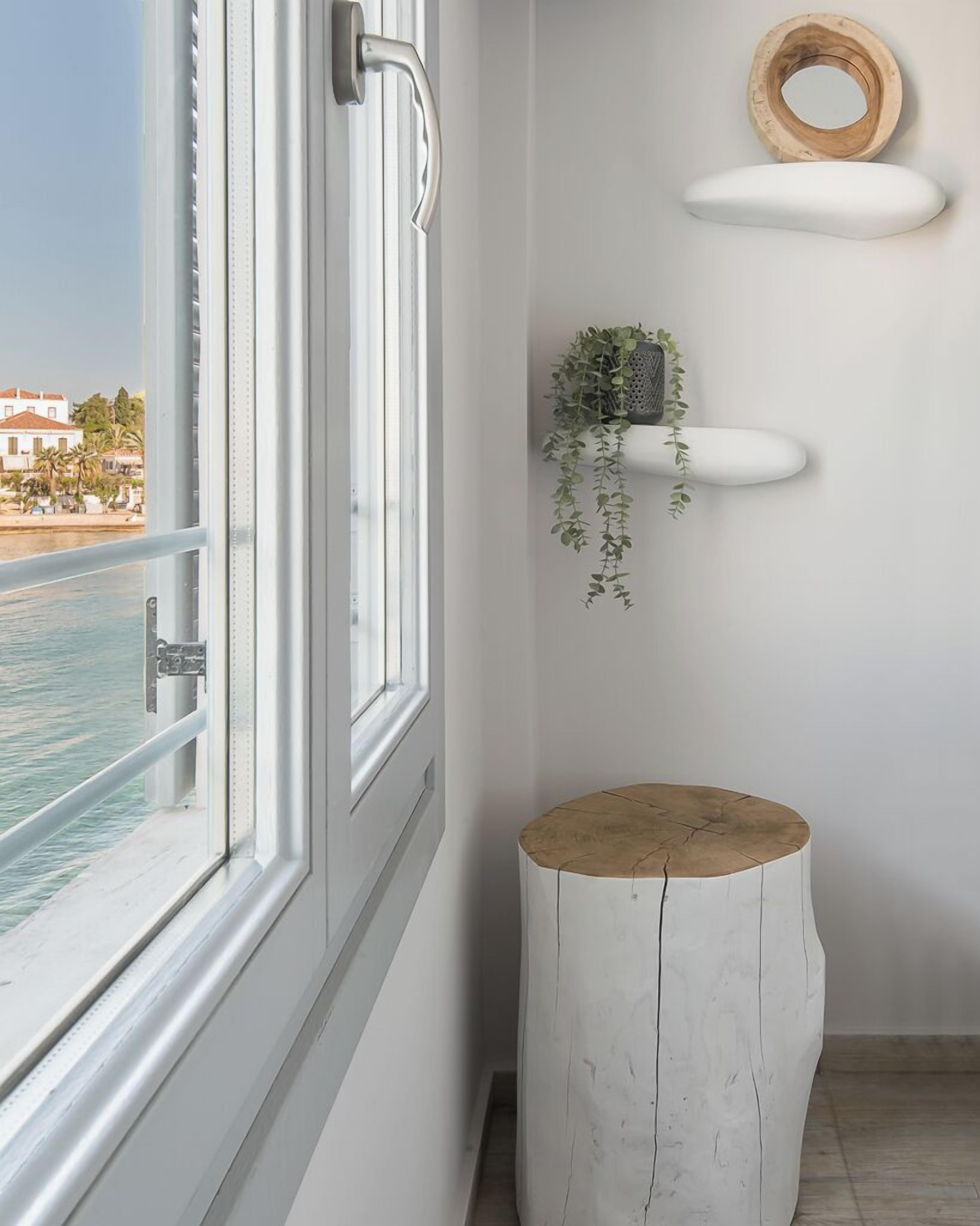 Una habitación con un gran ventanal con vistas a un cuerpo de agua, un taburete de madera y una estantería con una planta.