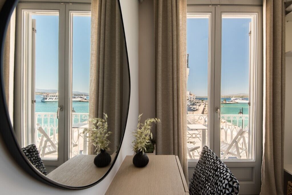 Habitación con balcón que da a un puerto deportivo y al mar. Se observa un espejo redondo en la pared, una mesita con un jarrón y una silla con un cojín estampado.