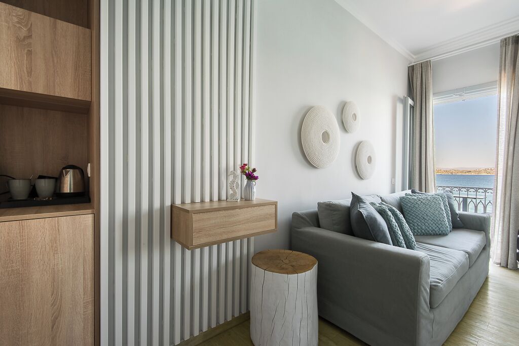 A modern living room with a grey sofa, wooden shelves, a small wooden stool, and a view of the sea through large windows.