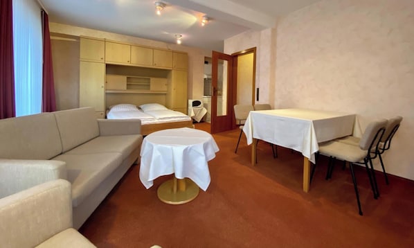 Habitación de hotel con sofá, una mesa redonda con mantel blanco, cama con ropa de cama blanca y una zona de kitchenette.