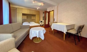Quarto de hotel com sofá, mesa redonda pequena, mesa de jantar com cadeiras, cama e cozinha compacta.