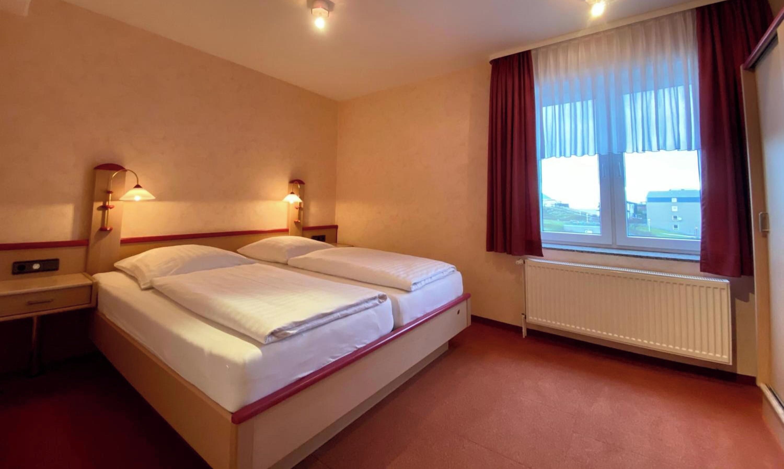 Ein Hotelzimmer mit Doppelbett, zwei Nachttischlampen, einem Fenster mit Vorhängen und einer Heizung.
