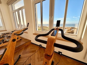 A home gym with exercise equipment and large windows overlooking a beach.