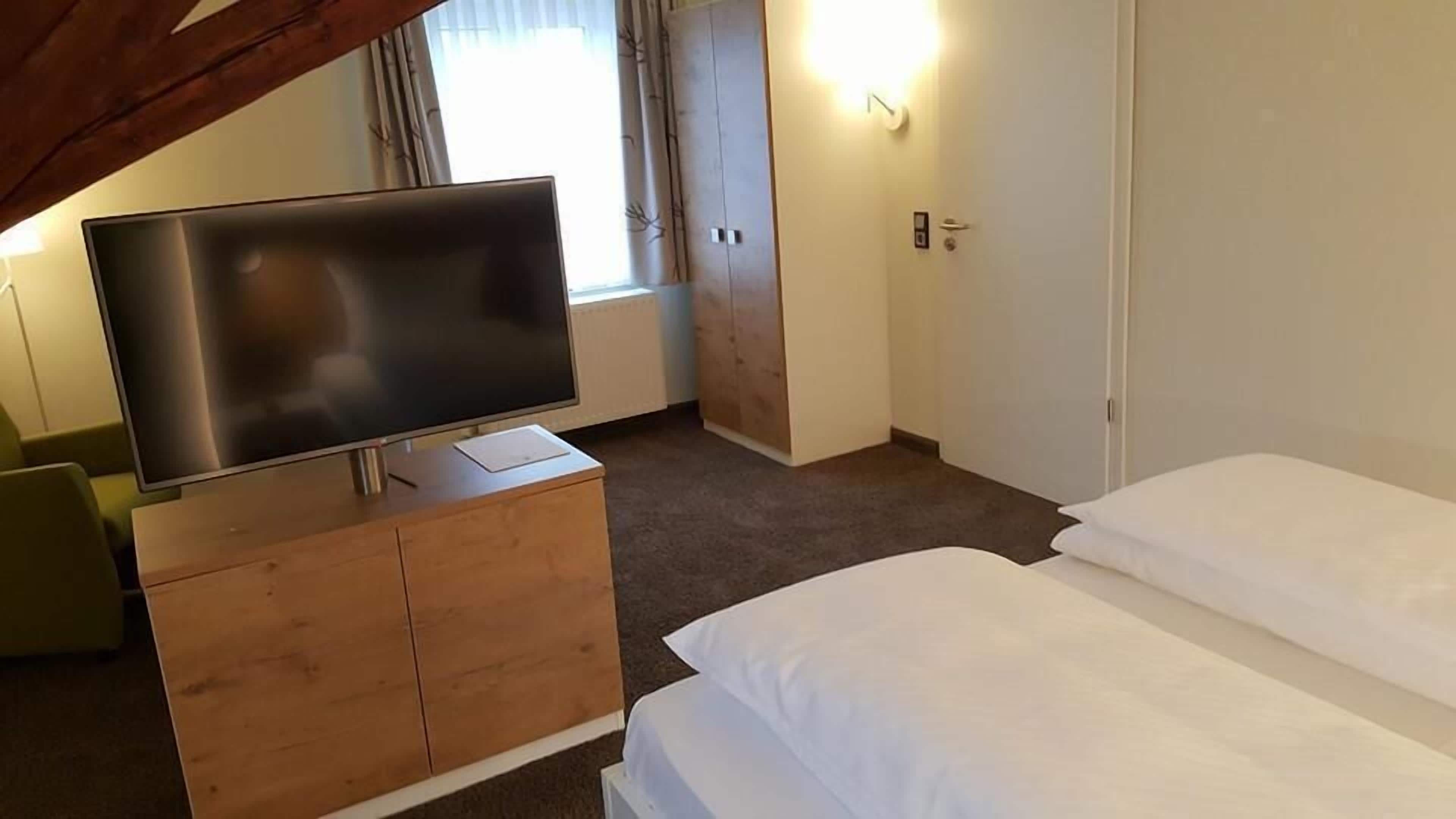 Habitación de hotel con televisor de pantalla plana sobre una cómoda de madera, cama con sábanas blancas, puerta con interruptor de luz y ventana con cortinas.