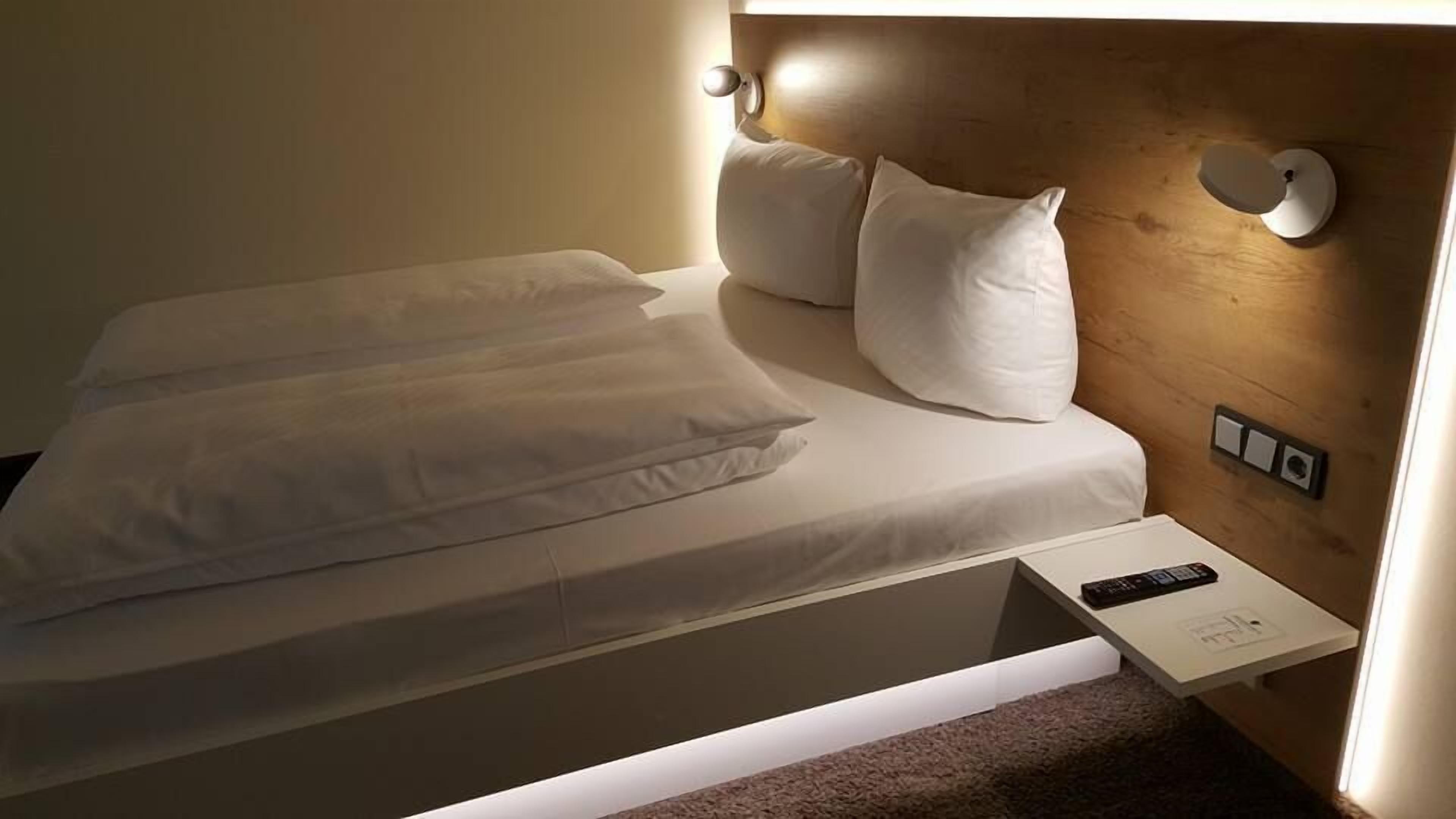 A neatly made bed with white linens and pillows, a bedside table with a remote control, and a wall-mounted light fixture.