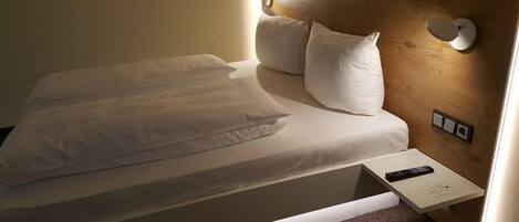 A neatly made bed with white linens and pillows, a bedside table with a remote control, and a wall-mounted light fixture.