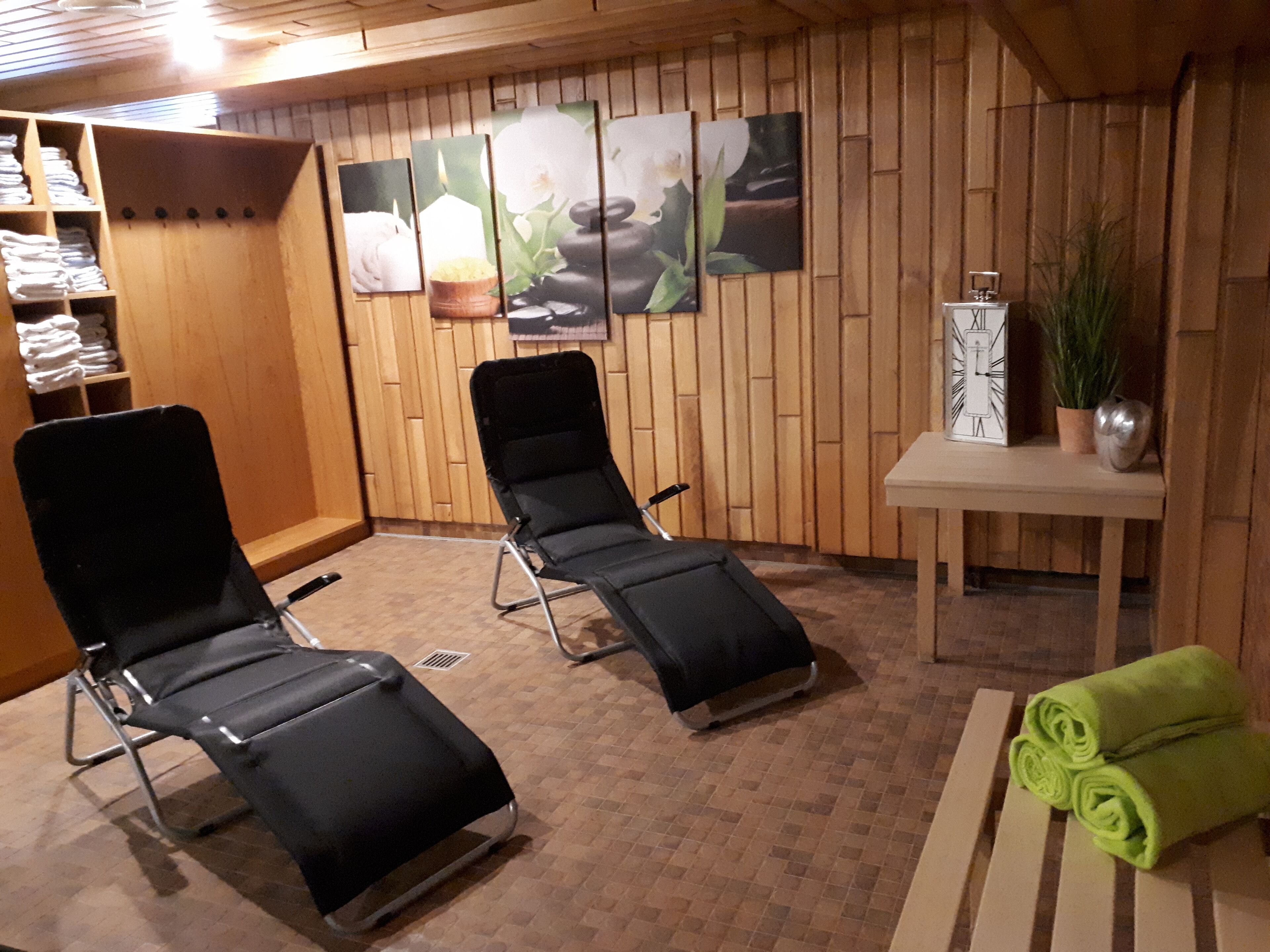 A spa room with two black massage chairs, a wooden wall with artwork, a small table with a plant, and neatly folded towels.