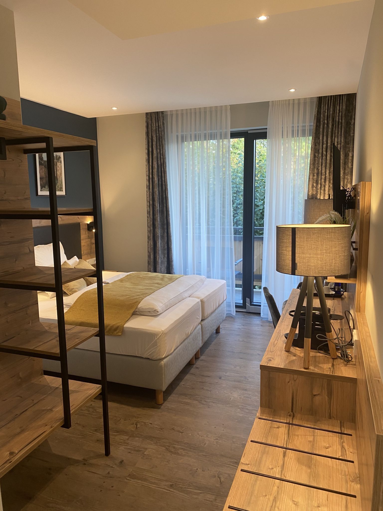 A modern bedroom with a bunk bed, a desk with a lamp, and a large window with sheer curtains.