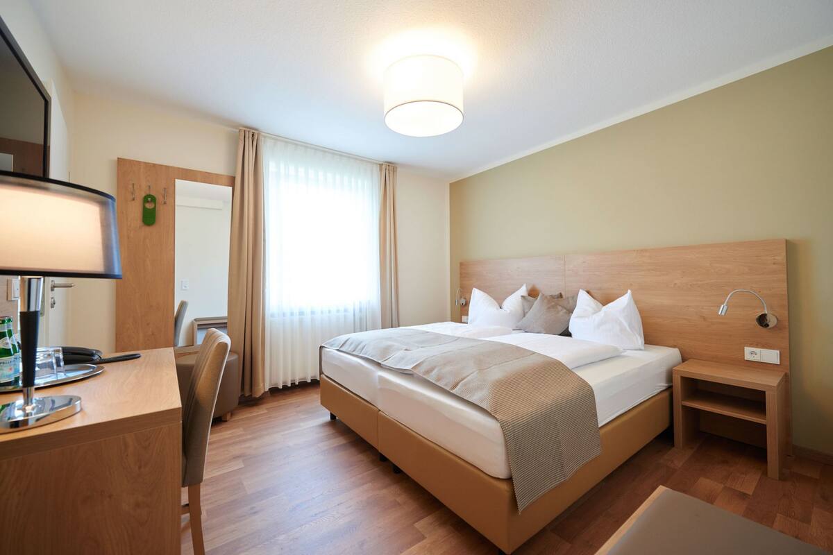 Une chambre d’hôtel comprenant un lit, une table de chevet, une lampe, un bureau, une chaise et une fenêtre avec des rideaux.