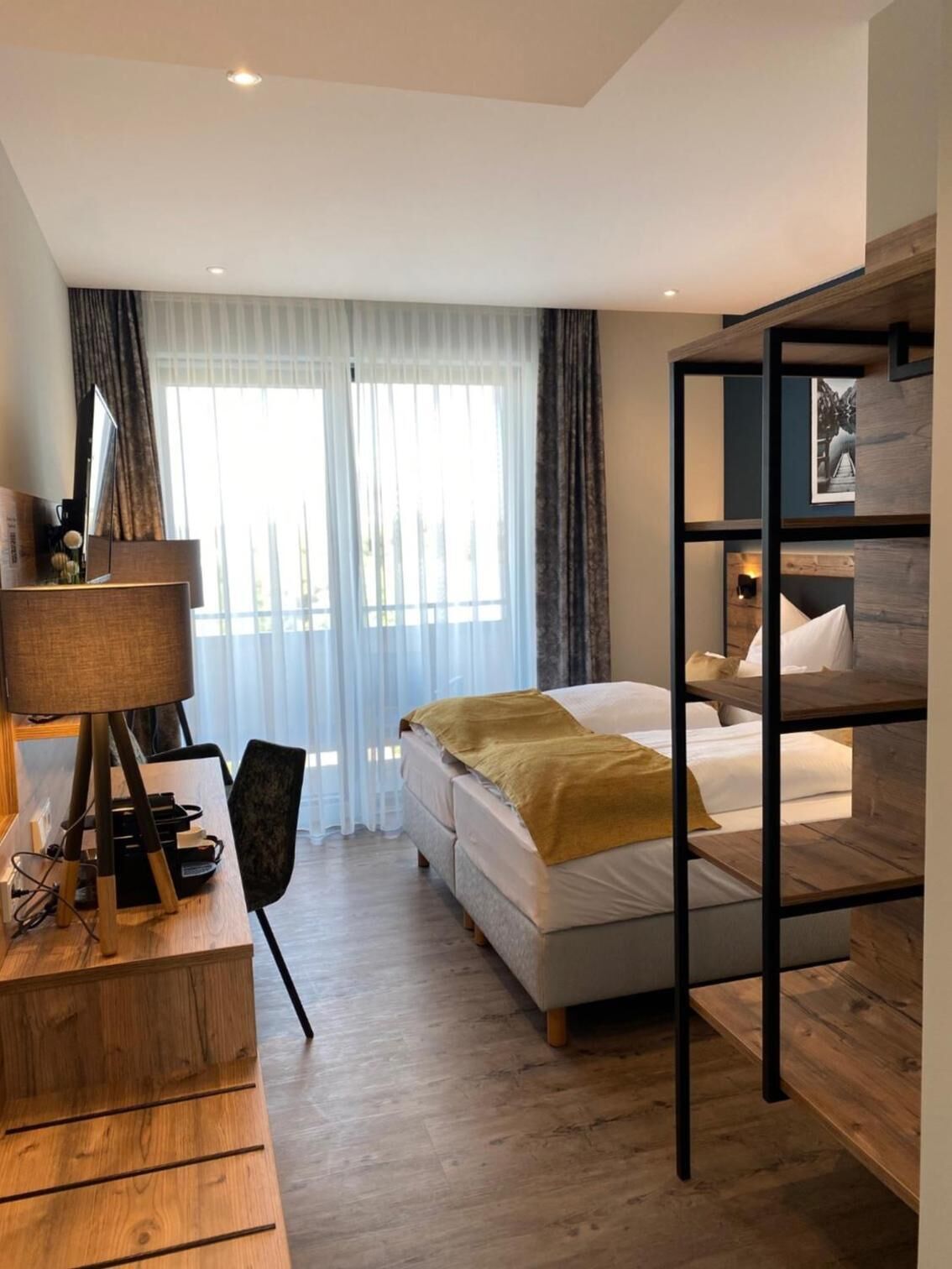 A modern hotel room with a bed, a desk with a lamp, a wooden shelf unit, and a large window with sheer curtains.