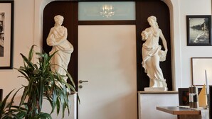 Una zona de recepción con dos estatuas blancas, un candelabro, cuadros enmarcados y una planta en maceta.