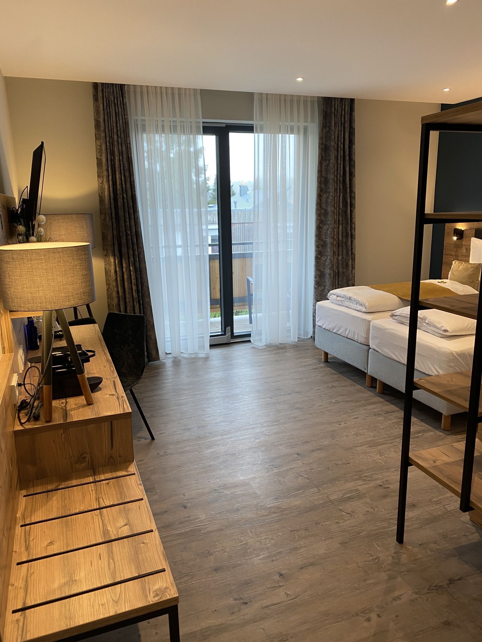 A hotel room with a wooden desk, a bed, and a view through a large window.
