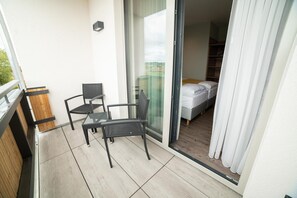 A balcony with two chairs and a table, a sliding glass door leading to a bedroom with a bed and a wardrobe.