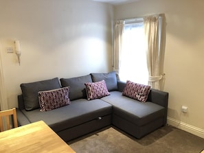A modern living room with a grey sectional sofa, patterned throw pillows, a wooden dining table, and a window with curtains. - North Circular Studios 5 (Dublin)