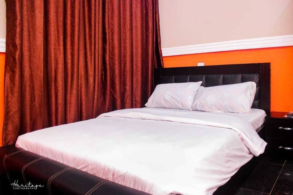 Uma cama com roupa de cama branca, cabeceira preta e cortina marrom.