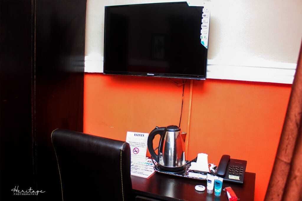 A hotel room with a television mounted on the wall, a coffee maker on the table, a telephone, and a sign indicating the room number.