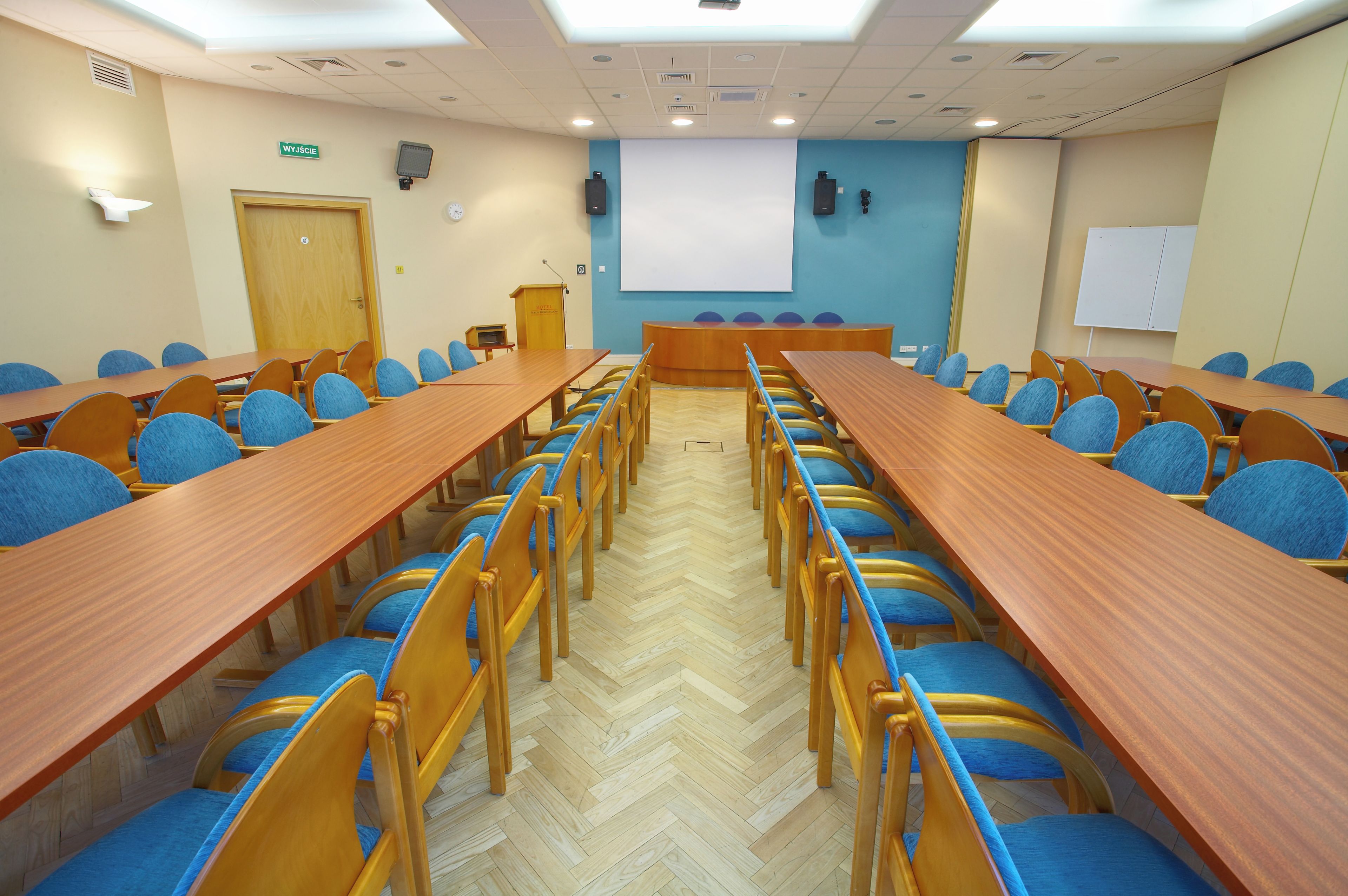 A conference room with long tables and chairs, a podium, a projector screen, and a clock on the wall.