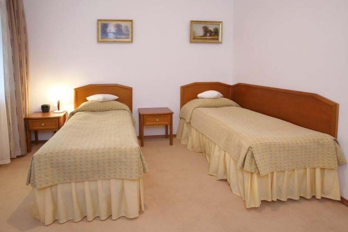 Habitación doble con dos camas individuales, una mesita de noche con lámpara y dos cuadros en la pared.