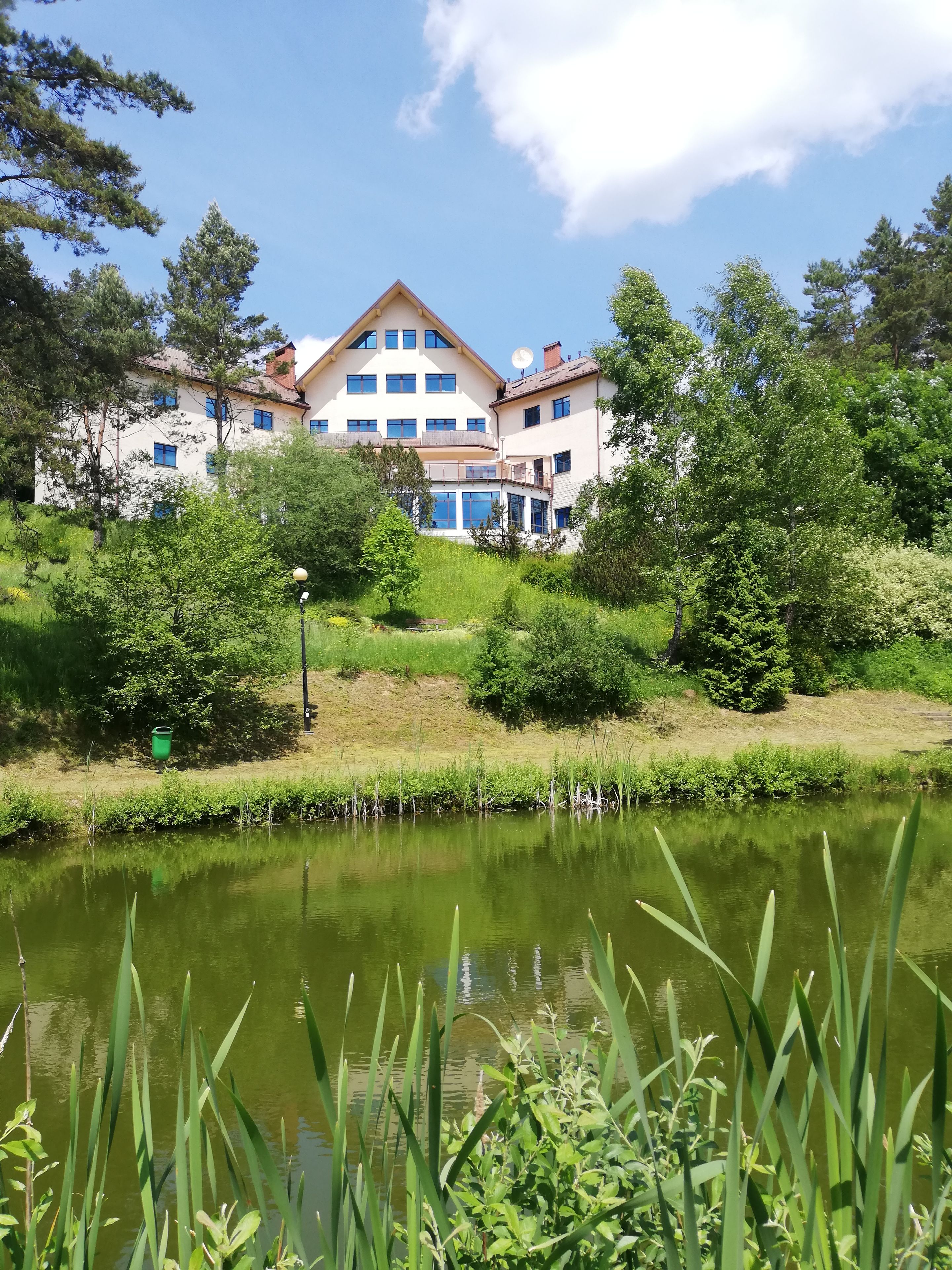 A large two-story building with multiple windows and a satellite dish, surrounded by lush greenery and a pond.