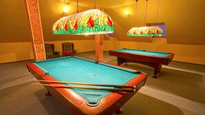Una mesa de billar con taco, otra mesa al fondo y una lámpara de vitral.