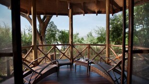 An outdoor wooden deck with wicker furniture and a table, surrounded by a wooden railing and a large tree trunk. - Jambo Impala Ecolodge (Kisumu)