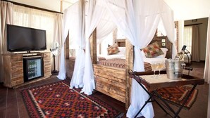 A room with a wooden bed, white canopy, a flat-screen TV, a minibar, and a patterned rug. - Jambo Impala Ecolodge (Kisumu)