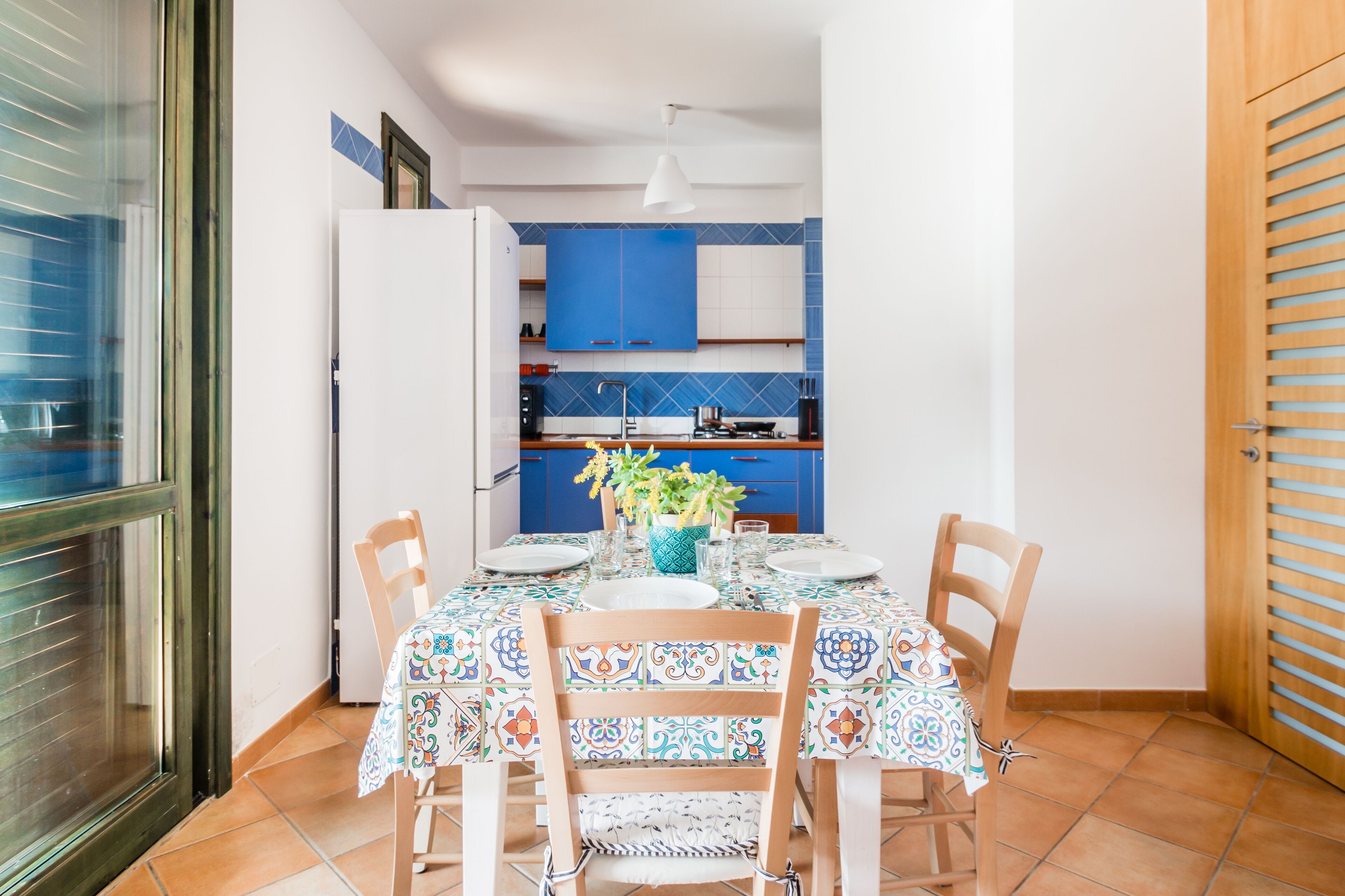 Un comedor con una mesa preparada para cuatro, una cocina azul al fondo y una puerta corredera de vidrio a la izquierda.