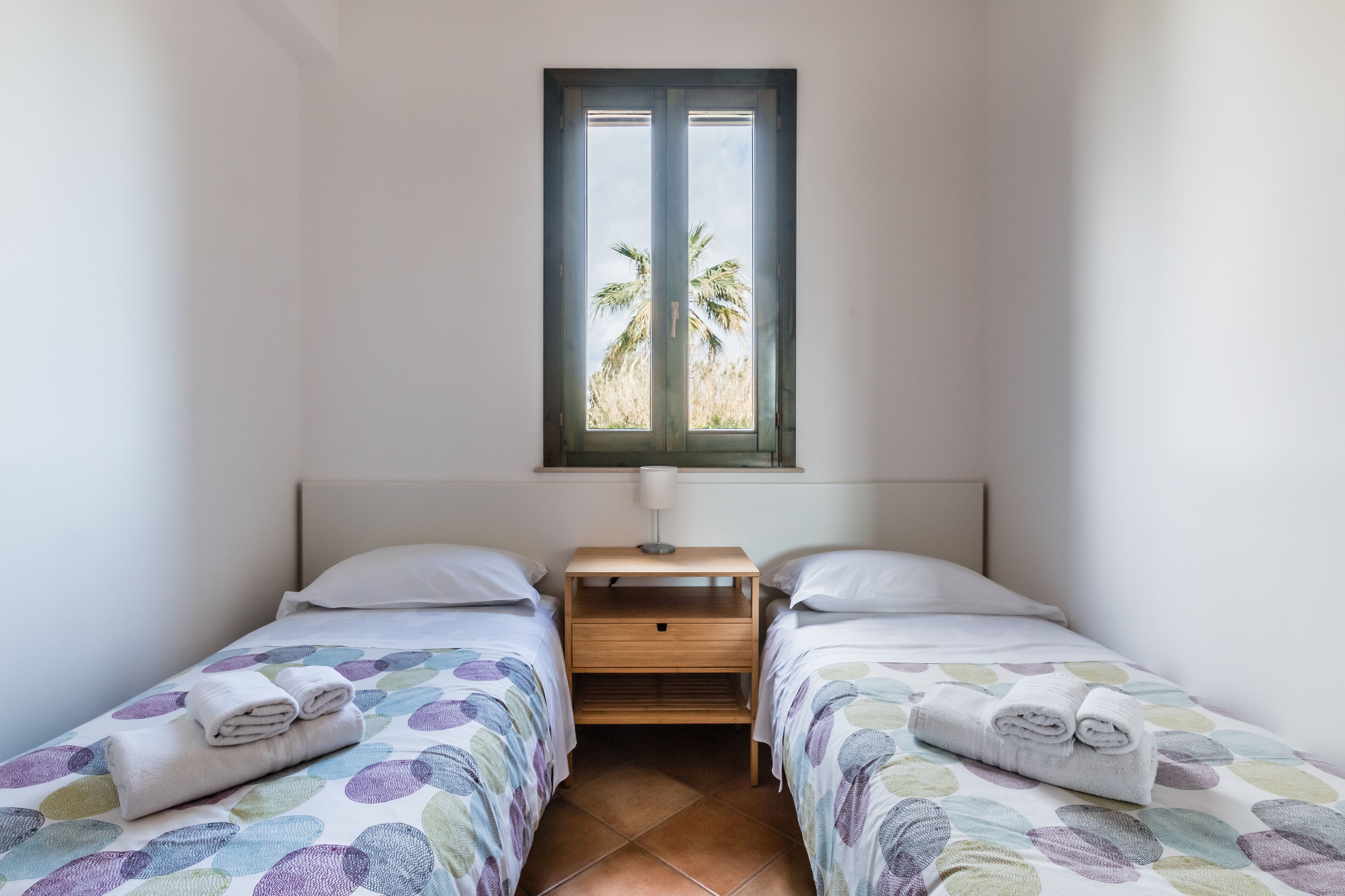 Een kleine, schone kamer met twee bedden, een raam met uitzicht op palmbomen en een nachtkastje met een lamp.
