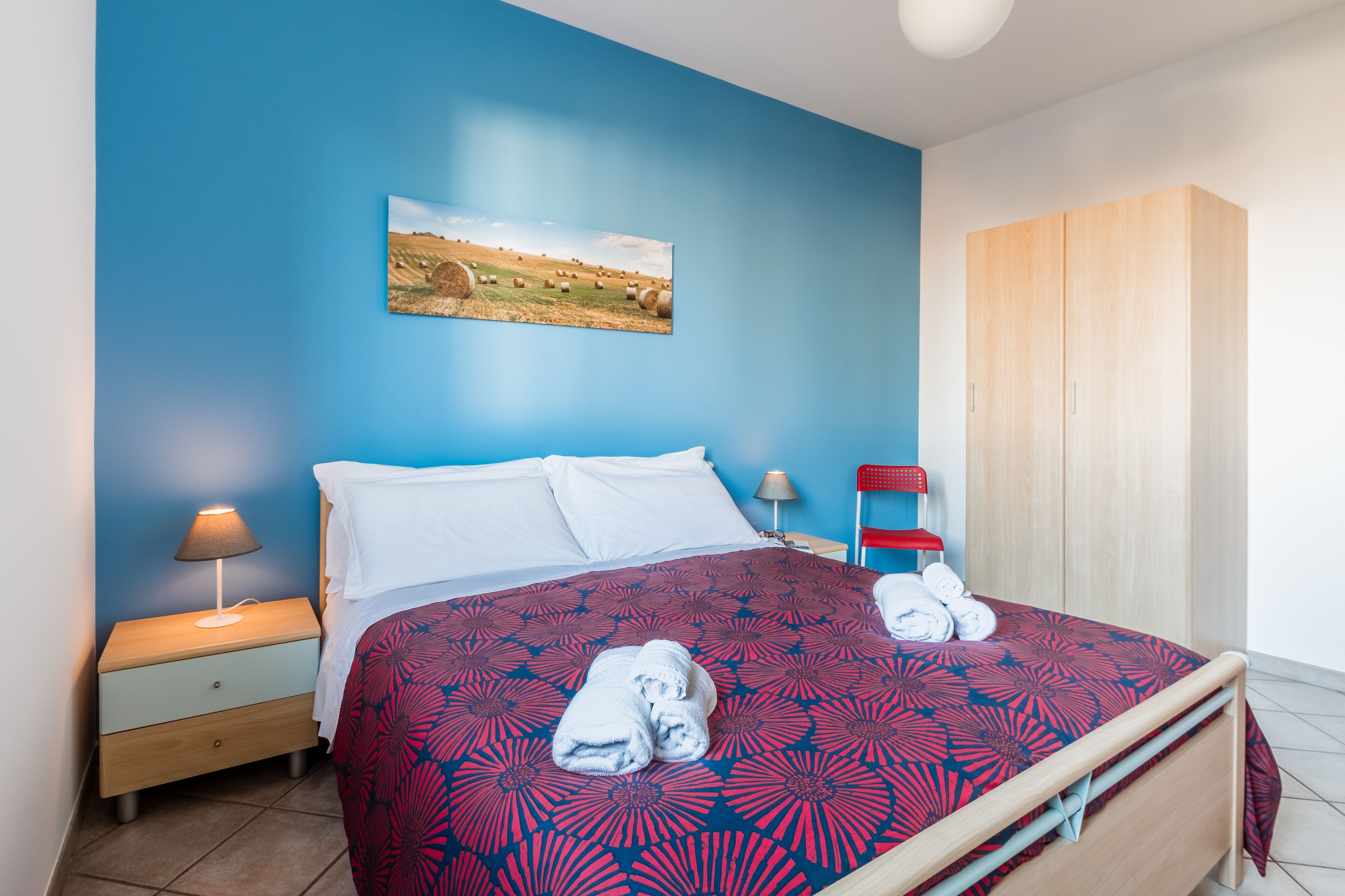 A neatly made bed with a patterned bedspread, a nightstand with a lamp, a red chair, and a wooden wardrobe in a room with blue walls.