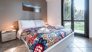 A modern bedroom with a large bed, a nightstand, a painting on the wall, and a window with a view of greenery. - Residence Andrea Doria (Ragusa)