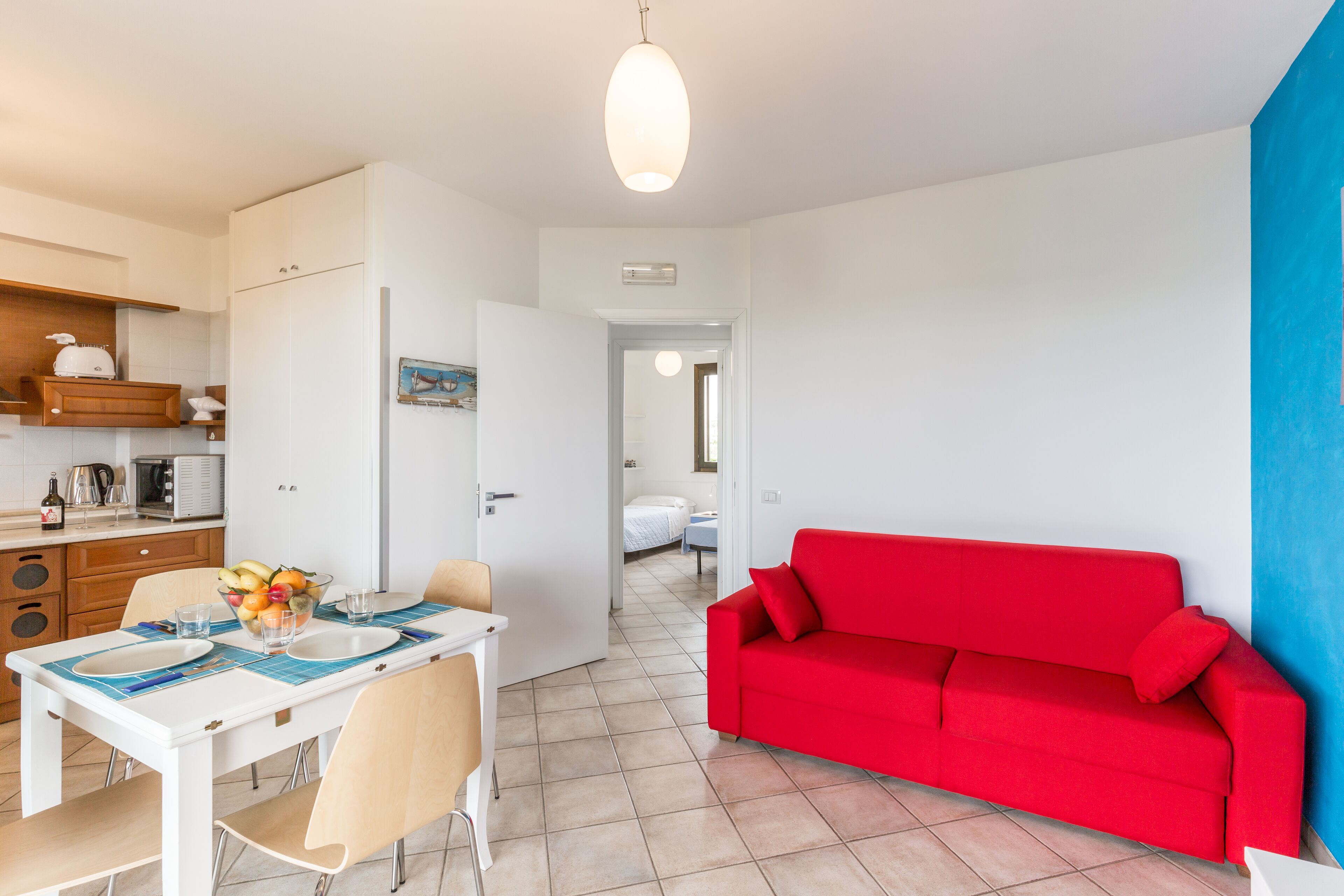A modern kitchen with white cabinets, a dining table set with plates and glasses, and a red sofa in a living area.