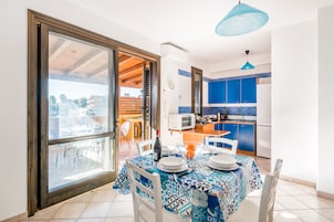 Un comedor con una mesa dispuesta para dos personas, una cocina al fondo y un ventanal con vistas al exterior.