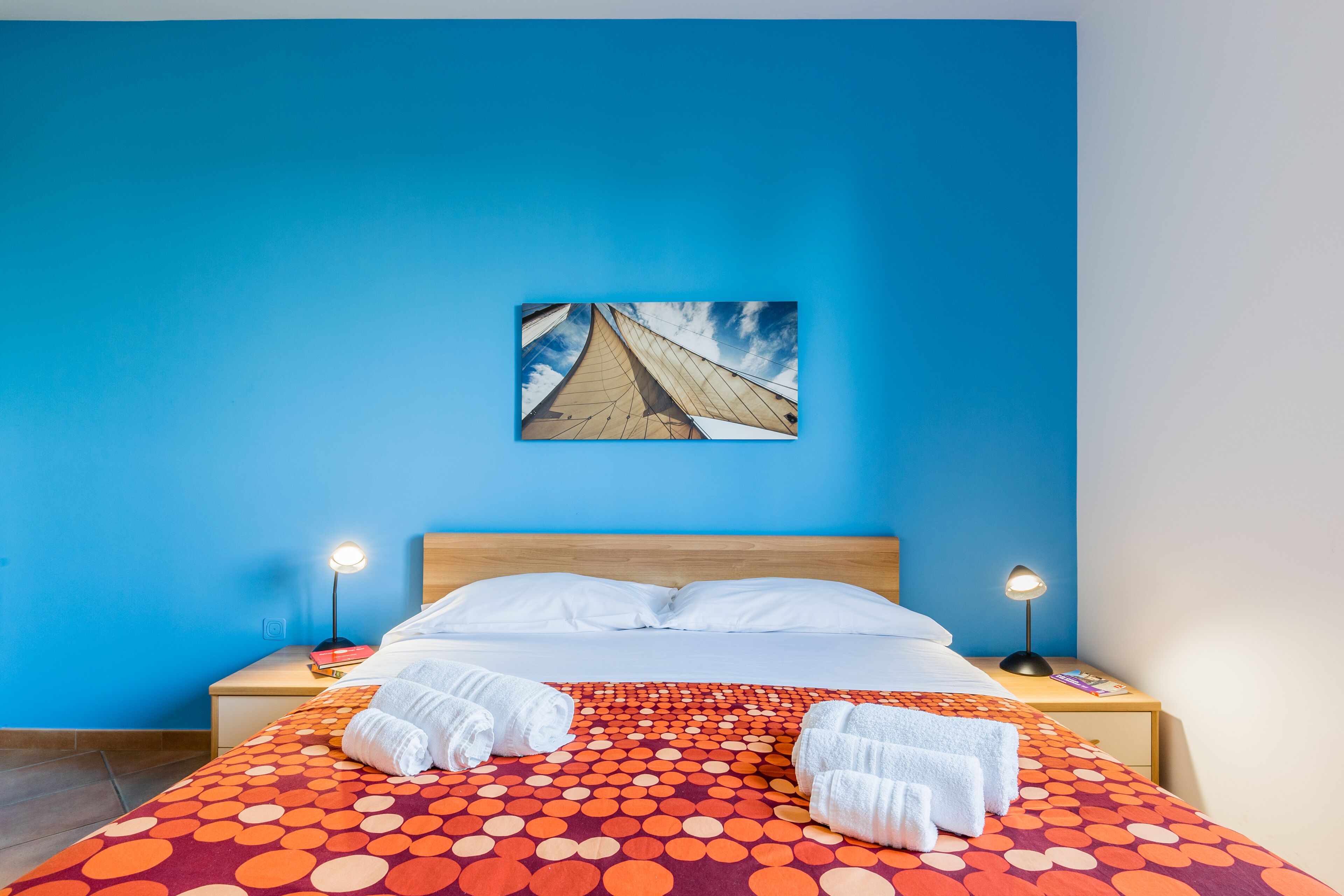 A hotel room with a blue wall, a bed with a polka dot bedspread, two white pillows, two bedside lamps, and a framed picture of a sailboat on the wall.