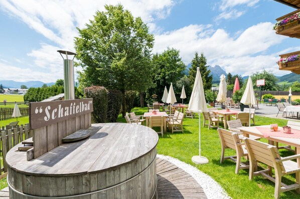 An outdoor seating area with wooden tables and chairs, surrounded by greenery and a sign reading 'Schattenbad'.
